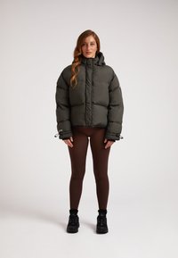 Giacca a vento verde scuro con colletto alto, dotata di polsini elastici e vestibilità rilassata, indossata sopra leggings marroni e scarpe nere.