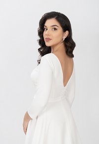 Weißes langärmeliges Kleid mit tief ausgesetztem Rücken, glattem Stoff, figurbetontem Oberteil, ausgestelltem Rock und dekorativen Knopfdetails auf der Rückseite.