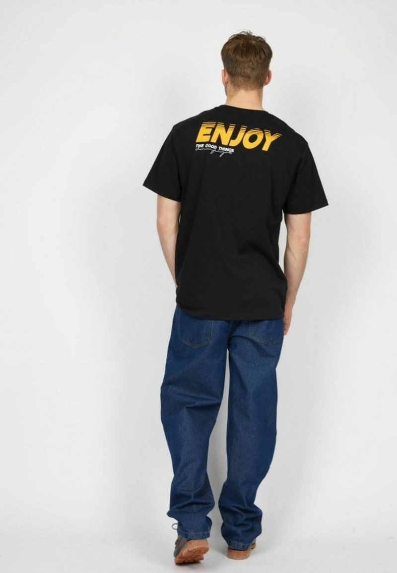 Sort bomulds T-shirt med gult tekst "ENJOY" på ryggen, parret med løse blå denim jeans og brune fodtøj. Enkel og afslappet design.