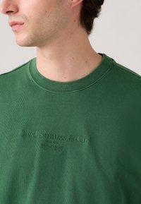 Zelené bavlněné tričko s kulatým výstřihem a reliéfním nápisem "Levi Strauss & Co." na přední straně. Hladký povrch a volný střih.