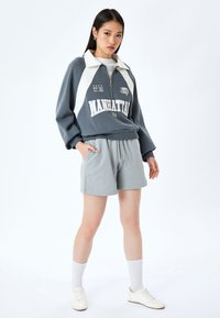 Felpa con zip grigia e bianca con colletto, caratterizzata da ricami "MANHATTAN USA" e stelle. Abbinata a pantaloni corti grigi chiari e sneakers bianche.