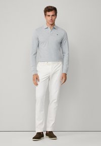 Harmaa pitkähihaisen poolopaidan kauluksella, yhdistettynä valkoisiin slim-fit-housuihin ja ruskeisiin slip-on-kenkiin, yksivärisen taustan edessä.