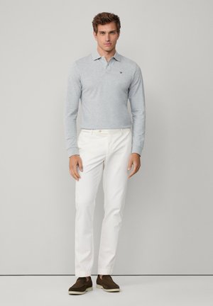 Szara koszulka polo z długim rękawem i kołnierzykiem, zestawiona z białymi spodniami o dopasowanym kroju oraz brązowymi butami wsuwanymi, na jednolitym tle.