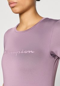 Μωβ αθλητική μπλούζα με στρογγυλή λαιμόκοψη και λεία υφή, με το λογότυπο "Champion" σε φωτεινότερη απόχρωση στο στήθος.