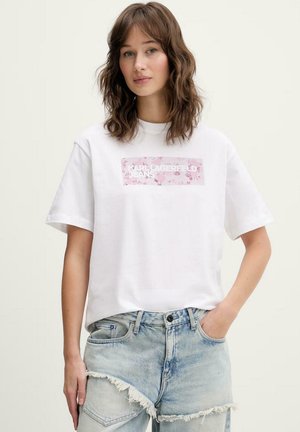 Giovane donna con capelli castani mossi che indossa una t-shirt bianca con logo floreale Karl Lagerfeld e pantaloncini di jeans chiari sfrangiati, mano in tasca.