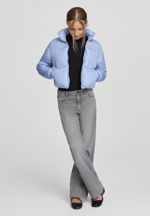 Giacca a vento azzurra chiara con colletto alto, polsini elasticizzati, abbinata a jeans dritti grigi e scarpe slip-on nere.