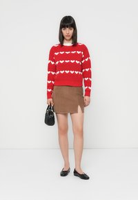 Czerwony sweter z dzianiny z białym wzorem serca, zestawiony z brązową zamszową spódniczką mini i czarnymi balerinami. Trzyma małą czarną torebkę.