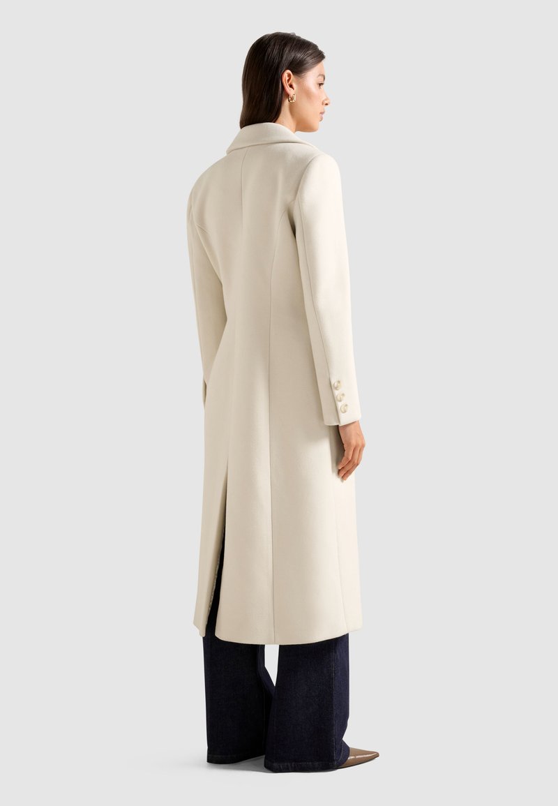 Cappotto lungo in lana color crema con un design aderente, colletto a punta e fessure laterali. Presenta dettagli con bottoni sulle maniche ed è abbinato a jeans scuri.