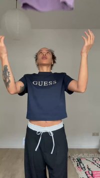 Osoba v tmavě modrém tričku s nápisem "GUESS" a černých kalhotách hází fialovou látku vzhůru v minimálně zařízené místnosti s vzorovaným kobercem.