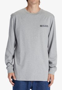 Long-sleeve gray cotton t-shirt featuring a black logo on the upper left chest and a classic crew neckline. Soft texture, relaxed fit.