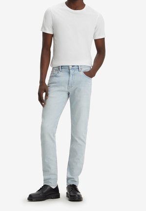 Mann trägt ein schlichtes weißes Kurzarmshirt, hellblaue Slim-Fit-Jeans und schwarze Schnürschuhe, steht mit einer Hand in der Tasche.