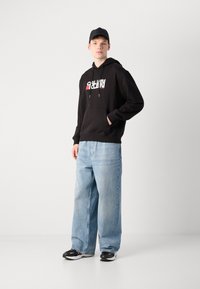 Svart hettegenser med hvit og rød grafikk, frontlomme. Lyseblå vide jeans. Svarte joggesko med hvite detaljer. Svart caps.
