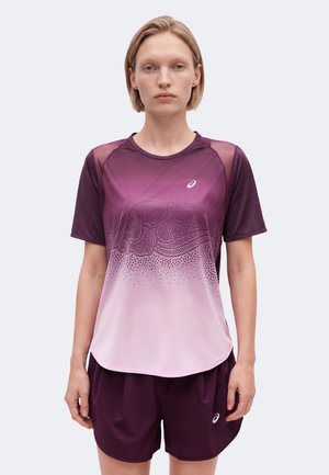 Femme debout portant un t-shirt de sport dégradé violet avec des détails à motifs et un short de sport violet assorti sur un fond uni.