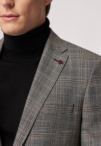 Blazer a quadri grigi con righe marroni e nere, vestibilità su misura, abbinato a un dolcevita nero. Presenta un piccolo accento rosso sulla giacca.