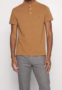Polo marron avec col, patte de boutonnage à trois boutons et manches courtes. Présente un petit logo sur la poitrine et une texture de tissu lisse.