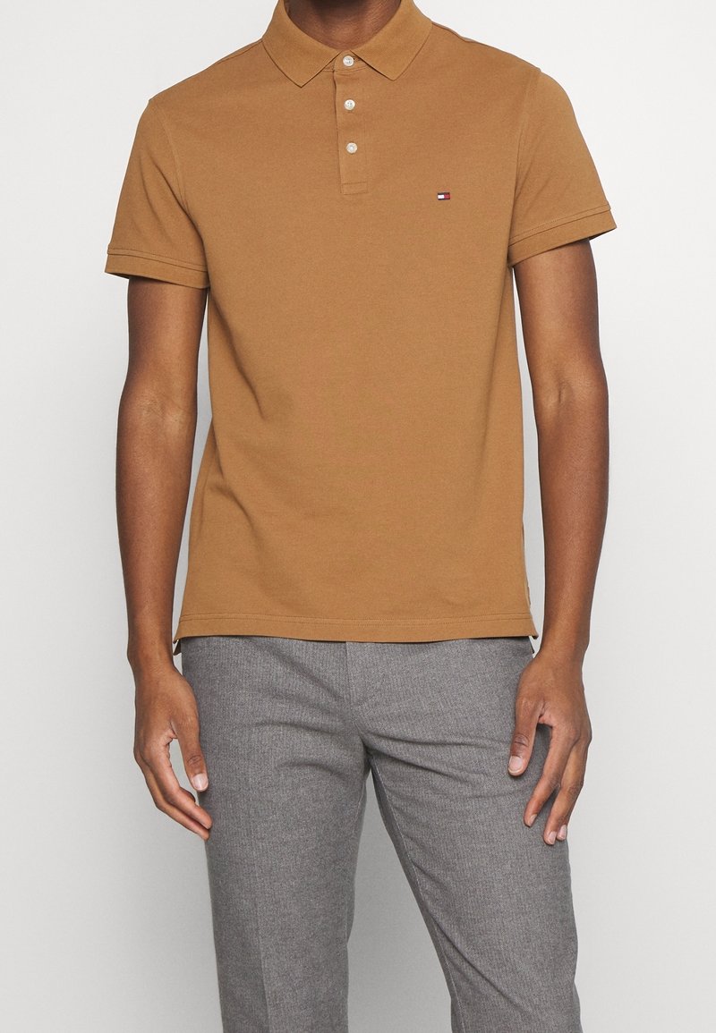Polo marron avec col, patte de boutonnage à trois boutons et manches courtes. Présente un petit logo sur la poitrine et une texture de tissu lisse.