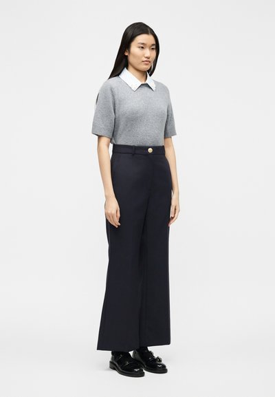 Femme debout portant un pull gris à manches courtes sur une chemise blanche à col, un pantalon ample marine et des mocassins noirs sur un fond blanc.