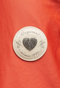Emblema metálico en forma de corazón sobre un parche de cuero blanco, cosido con "Raqueur desde 1997", situado sobre un fondo de tela naranja.