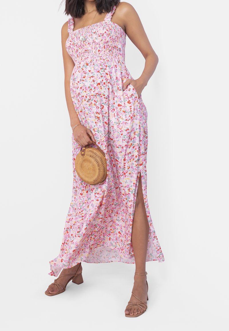 Robe maxi florale en rose pastel avec un corsage smocké, des bretelles fines, une fente sur le côté et un tissu léger et fluide. Accessoire : sac rond en paille.