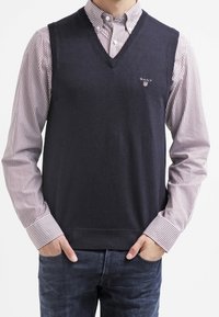 Marineblauer Strickweste mit V-Ausschnitt, darüber getragen ein gestreiftes weiß-rotes Hemd mit Kragen, mit kleinem Logo auf der linken Brust.