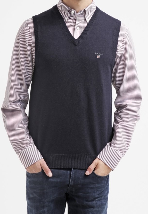 Marineblauer Strickweste mit V-Ausschnitt, darüber getragen ein gestreiftes weiß-rotes Hemd mit Kragen, mit kleinem Logo auf der linken Brust.