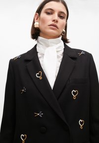 Blazer nero su misura con vari dettagli decorativi, tra cui spille a forma di fiore e cuore. Camicetta bianca fresca con colletto a fiocco.