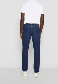 Vaqueros azul oscuro de mezclilla, con bolsillos traseros con detalles de costura, combinados con una camiseta blanca lisa y deportivas claras.