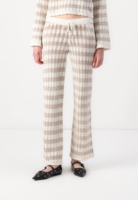 Pantalon de lounge en tricot rayé beige et blanc, avec une taille à cordon de serrage, une forme évasée et une texture en tissage ouvert.