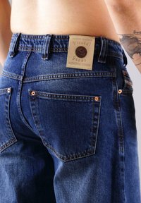 Nahaufnahme einer Person, die blaue Denim-Jeans mit einem Lederlabels ausgestellt "Picaldi Jeans" am Bund trägt und ein Tattoo am Arm hat.