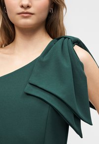 Mujer con vestido verde oscuro de un solo hombro, detalle grande de tela en capas en el hombro descubierto, pendiente de aro dorado visible.