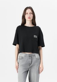 Schwarzes, kurzärmeliges, aus Baumwolle bestehendes Cropped-T-Shirt mit einer Brusttasche und einem Etikett, kombiniert mit grauen Jeans aus Denim.
