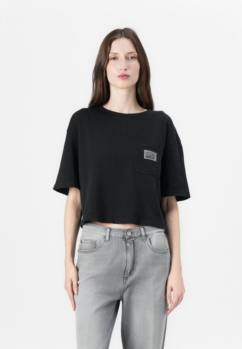 Schwarzes, kurzärmeliges, aus Baumwolle bestehendes Cropped-T-Shirt mit einer Brusttasche und einem Etikett, kombiniert mit grauen Jeans aus Denim.