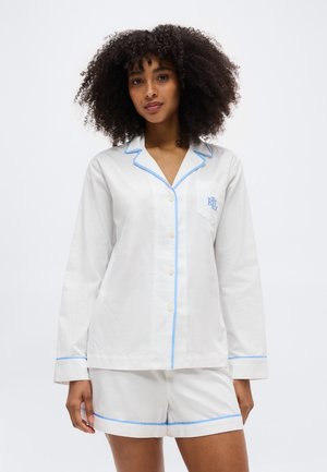 Vrouw met krullend haar, gekleed in een wit lang-sleeve overhemd met knopen en bijpassende korte broek met lichtblauwe bies en "RL" borduursel op het borstzakje.