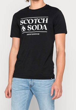 Person iført sort kortærmet t-shirt med hvid tekst "Scotch & Soda Amsterdam" og lyseblå jeans, stående med afslappede arme.