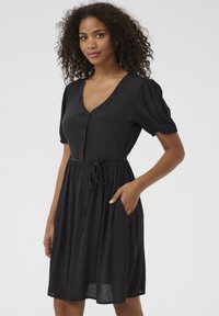 Robe noire à manches courtes avec un décolleté en V, un bouton à l'avant et une taille à nouer. Présente un design fluide et des poches latérales. Tissu doux et léger.