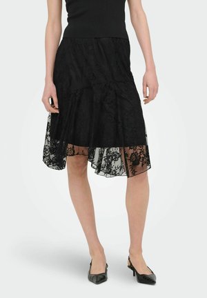 Jupe en dentelle noire à ourlet asymétrique portée avec un top sans manches noir et des escarpins slingback noirs à bout pointu sur fond blanc.