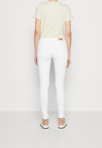 Jeans blancos ajustados con una textura suave, que presentan una cintura de altura media, dos bolsillos traseros y un parche de cuero marrón en la cintura.