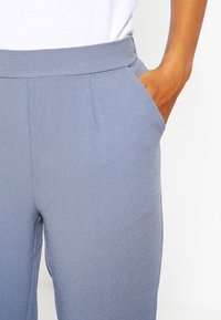 Grå, texturerade byxor med platt midja och sidofickor, delvis visade med en hand vilande i en ficka.