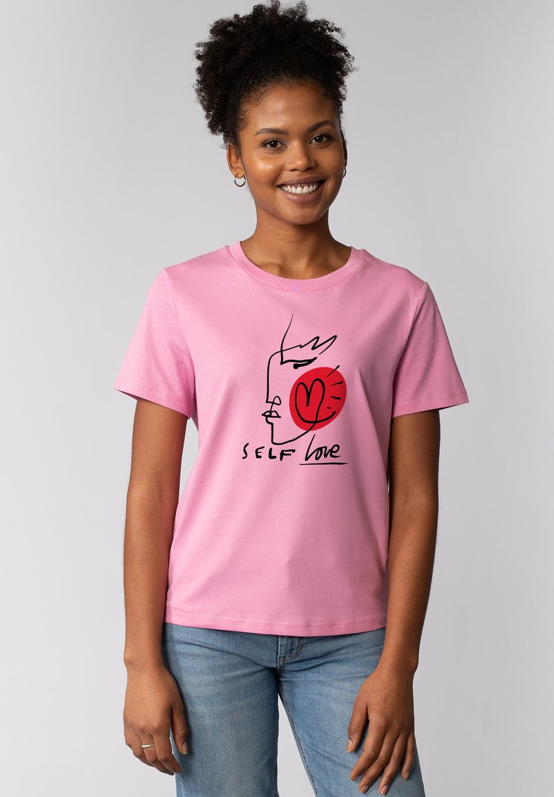 Roze katoenen T-shirt met een zwarte lijntekening van een gezicht en een rode hart, met de tekst "SELF love" in een cursieve lettertype. Korte mouwen.