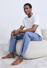 Hvit t-skjorte med korte ermer med sort "BOSS"-logo, kombinert med lyseblå bukser. Personen poserer på en kremfarget pute med tekstur.