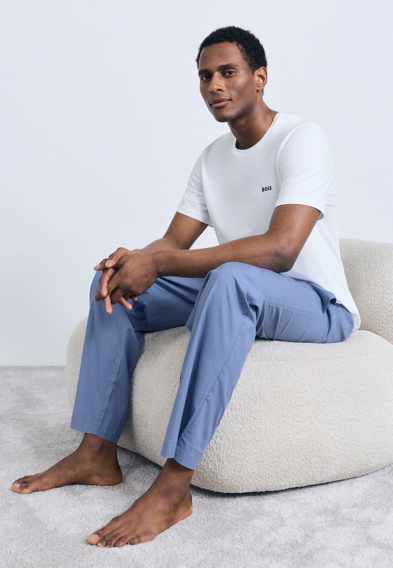 Hvit t-skjorte med korte ermer med sort "BOSS"-logo, kombinert med lyseblå bukser. Personen poserer på en kremfarget pute med tekstur.