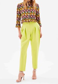 Mønstret bluse i flere farver med klokkeærmer, parret med lysegrønne plisserede bukser. Hvide strophæle fremhæver outfittet.