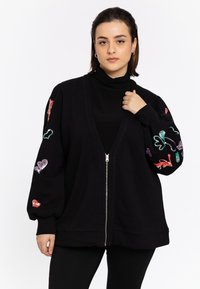 Zwarte cardigan met ritssluiting en een diepe V-hals, met kleurrijke, sprankelende hart- en dierenmotieven op de mouwen. Glad materiaal en een relaxte pasvorm.