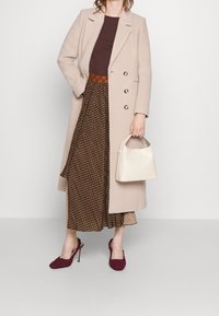 Beige wollen jas met dubbele knopen, bruin geribd topje, geblokte bruine rok, kastanjebruine hakken en een kleine witte handtas.