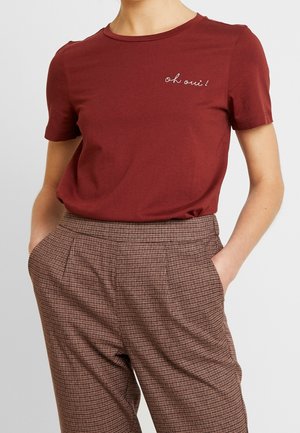 Osoba ubrana w bordową koszulkę z krótkim rękawem z napisem "oh oui!" oraz brązowe spodnie w kratę z wysokim stanem, z rękami w kieszeniach.