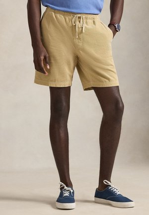 Mann trägt beige Shorts mit Kordelzug, blaue Sneakers mit weißen Sohlen und ein blaues Hemd, steht mit einer Hand in der Tasche.