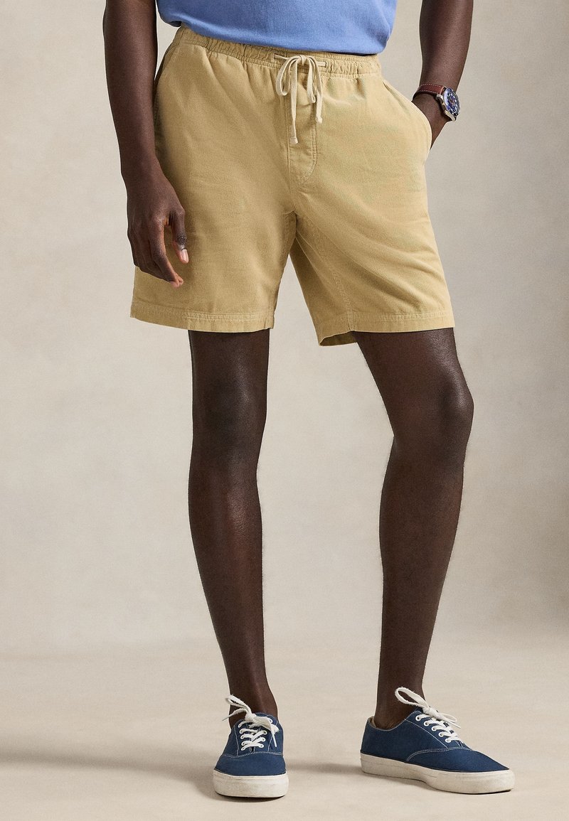 Homme portant un short beige à cordon, des baskets bleues avec semelles blanches et une chemise bleue, debout avec une main dans la poche.