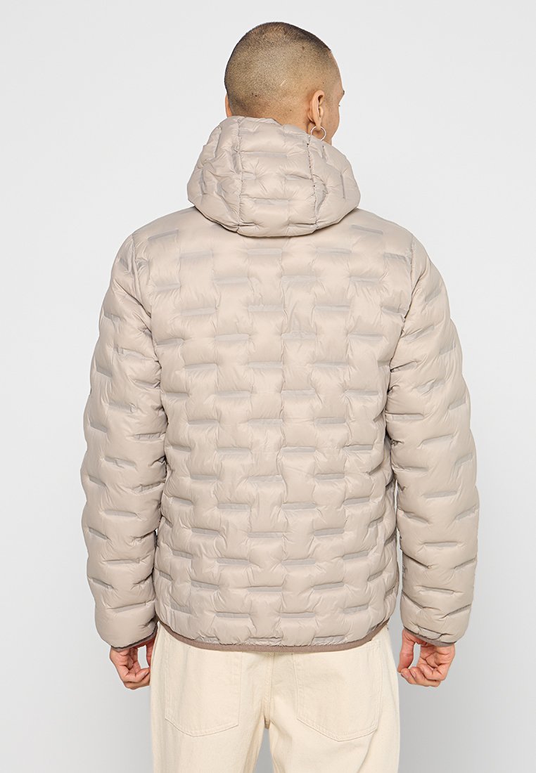 Mężczyzna w jasnobeżowej pikowanej kurtce puchowej z kapturem, zwrócony tyłem, z rękami swobodnie opartymi wzdłuż ciała, na jednolitym jasnym tle.