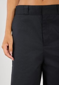 Pantalons noirs adaptés avec une texture lisse, présentant une taille plate, des passants de ceinture et une coupe classique, exposés avec une main reposant à proximité.