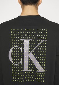 Personne portant un sweat-shirt noir avec un texte jaune répétitif "Calvin Klein Jeans Established 1978" et un grand logo gris "CK" dans le dos.